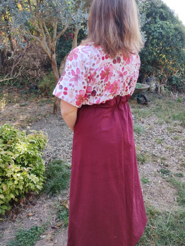 Vue de 3/4 dos d'une femme portant la robe longue manches courtes rose poudre et bas de robe uni bordeaux.
